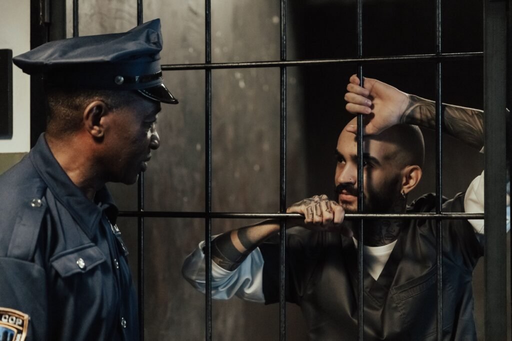 A police officer communicates with a tattooed prisoner behind bars in a dimly lit cell.